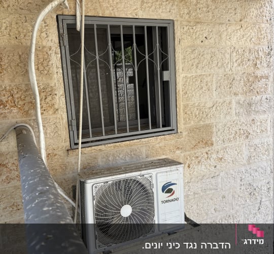 יחידת מיזוג אוויר מותקנת על קיר חיצוני עם חלון סורג.